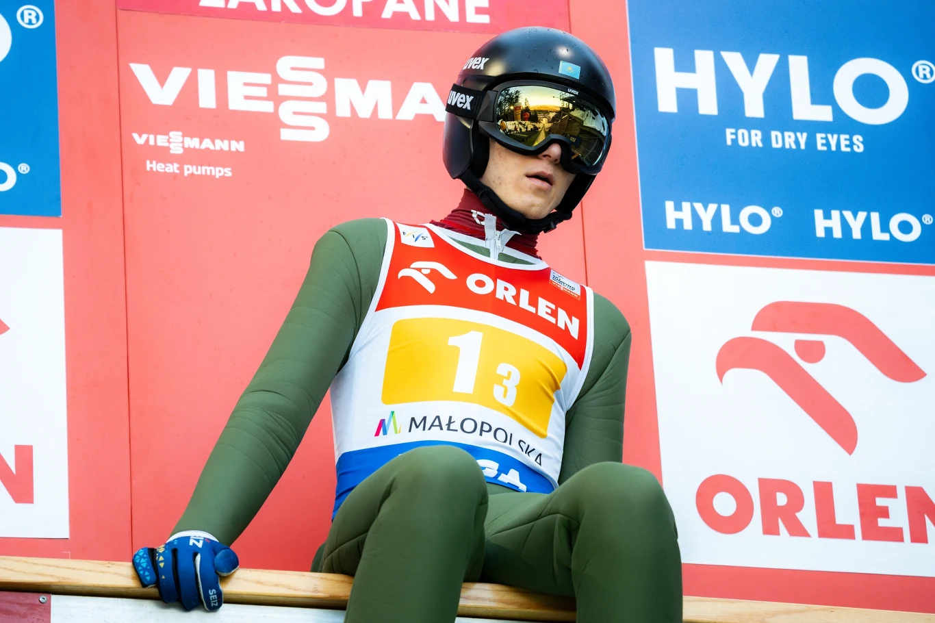 Ilja Miziernych Skoczek narciarski w zielonym kombinezonie i kasku z okularami przygotowuje się do skoku, siedząc na belce startowej przed ścianą z logotypami sponsorów.