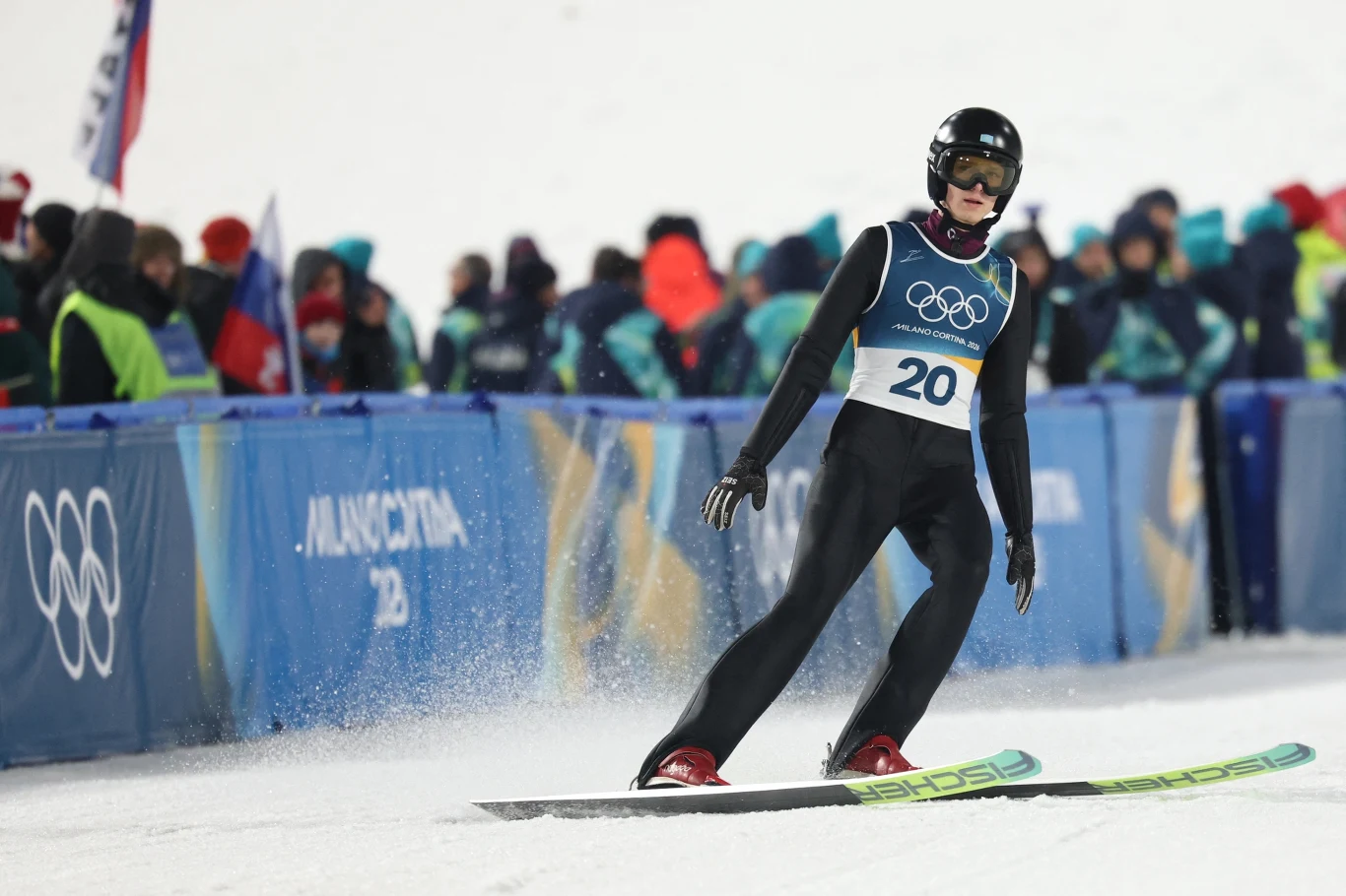 Ilja Miziernych Zawodnik ubrany w kombinezon narciarski i kask ląduje na nartach podczas konkursu skoków narciarskich na tle tłumu kibiców oraz banerów z logo igrzysk olimpijskich.