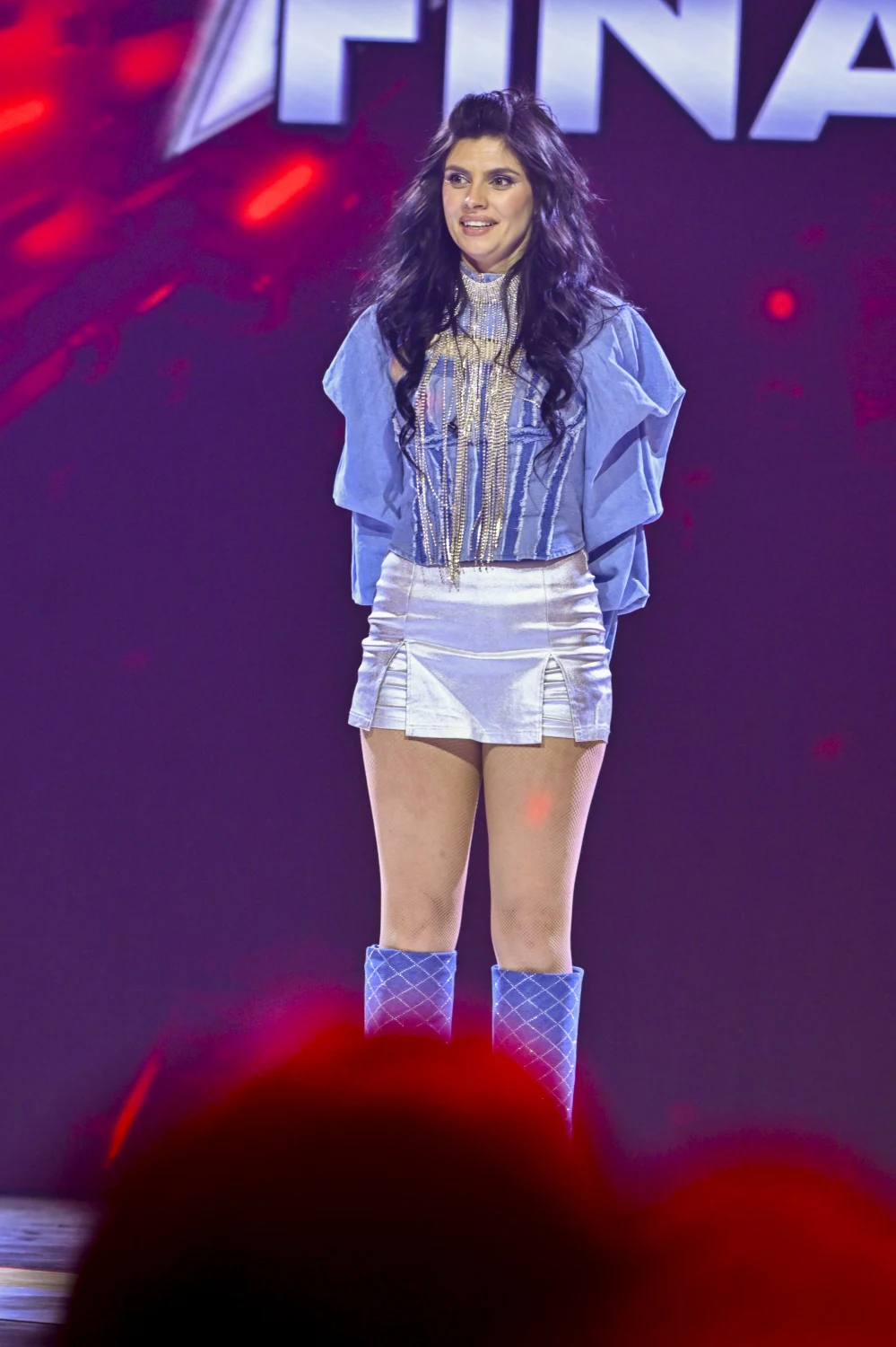 Młoda kobieta z ciemnymi, długimi włosami stoi na scenie podczas finału, ubrana w jasnoniebieską, błyszczącą stylizację z krótką spódnicą i wysokimi kozakami, w tle widoczne kolorowe reflektory i fragment napisu.