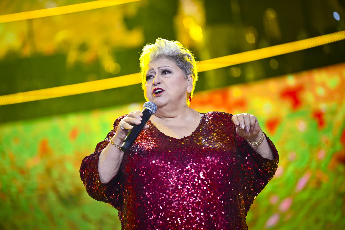 Kobieta śpiewająca na scenie w błyszczącej, czerwonej sukni, trzyma mikrofon, za nią kolorowe, rozświetlone tło.