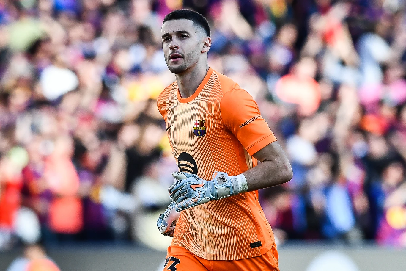 Bramkarz w pomarańczowym stroju FC Barcelony stoi na boisku piłkarskim, w tle rozmyta widownia na trybunach.