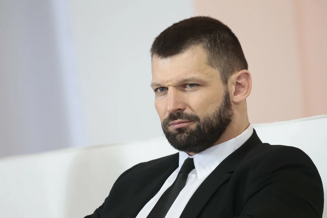Szymon Kołecki Mężczyzna z krótkimi włosami i brodą, ubrany w ciemny garnitur i białą koszulę, siedzi na białym tle z poważnym wyrazem twarzy.