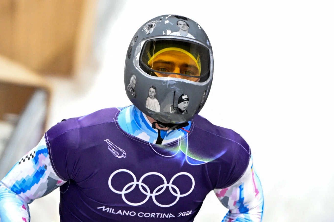 Sportowiec w stroju startowym oraz kasku ze zdjęciami postaci, ujęty na tle lodowego toru, trzymający głowę w pozycji skupienia. Na koszulce widoczne logo oraz napis Milano Cortina 2026, oznaczający zbliżające się zimowe igrzyska olimpijskie.