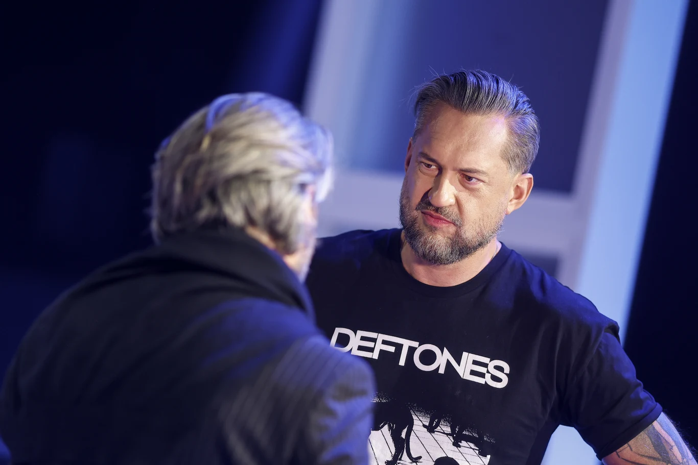 Marcin Prokop Dwóch mężczyzn rozmawia ze sobą, jeden z nich ubrany jest w czarną koszulkę z napisem Deftones i ma krótko przystrzyżoną brodę, drugi widoczny od tyłu, w ciemnej kurtce z siwiejącymi włosami.