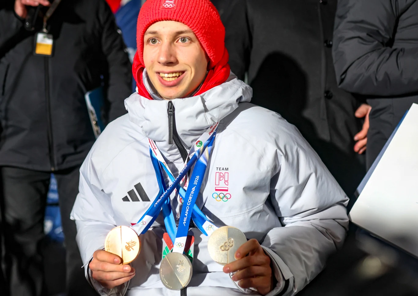 Młody sportowiec ubrany w białą kurtkę olimpijską oraz czerwoną czapkę trzyma trzy medale olimpijskie zawieszone na niebieskich wstążkach, uśmiecha się szeroko, w tle inni ludzie ubrani w ciemne kurtki.