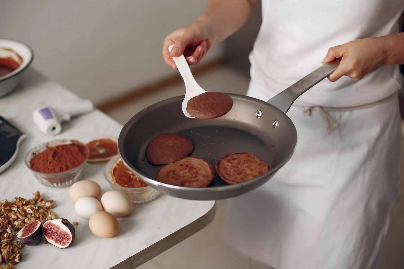 Osoba smaży czekoladowe placuszki na patelni, korzystając z łopatki kuchennej. Na stole obok znajdują się składniki potrzebne do przygotowania deseru: świeże figi, jajka, orzechy, kakao i miseczki z dodatkami.