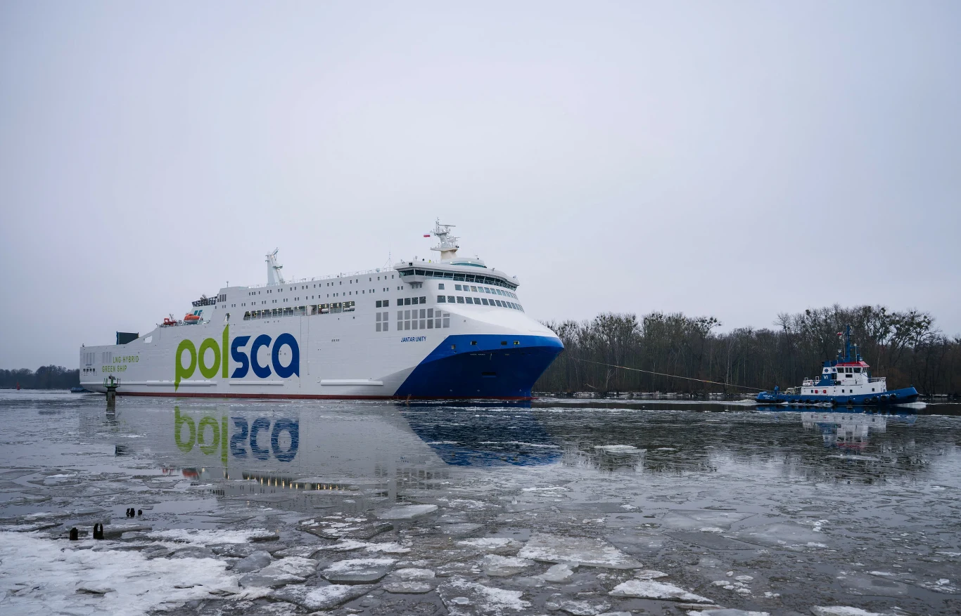 Jantar Unity to pierwszy od lat prom zbudowany w polskiej stoczni Prom pasażerski z logotypem Polferries na burcie stoi na częściowo zamarzniętej wodzie, obok znajduje się holownik, a w tle widoczne są drzewa pod pochmurnym niebem.