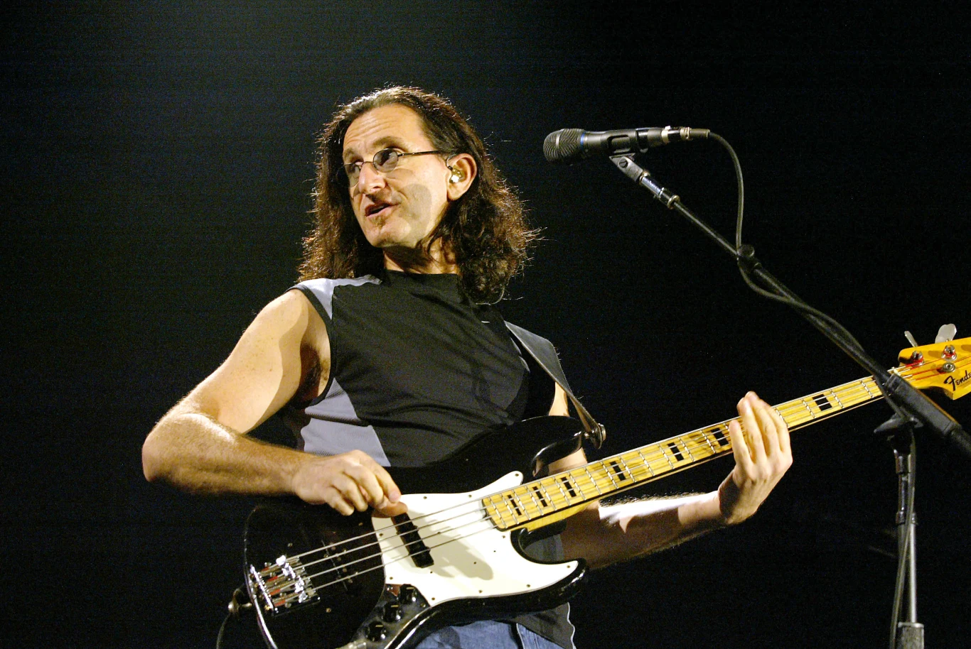 Geddy Lee (Rush) w 2004 r. Muzyk grający na gitarze basowej na scenie, z mikrofonem ustawionym przed nim, dynamiczne światło sceniczne podkreślające sylwetkę artysty.