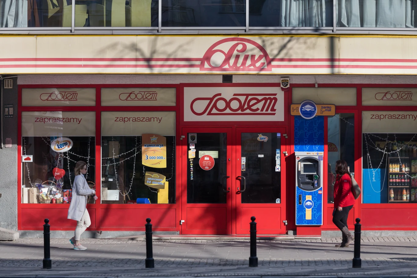 Te sklepy zna każdy Polak. Teraz została ich garstka Fasada sklepu Społem z czerwonymi drzwiami i witrynami, automat z napojami przy wejściu, dwie kobiety przechodzące chodnikiem przed sklepem, widoczne napisy zapraszające do środka oraz wystawione produkty w oknach.