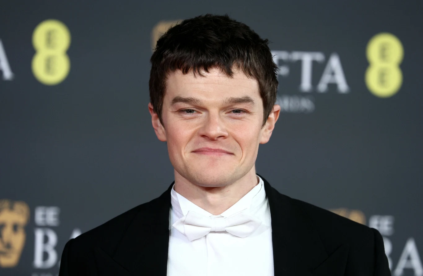 Robert Aramayo na 79. ceremonii rozdania nagród BAFTA Mężczyzna w smokingu i białej muszce stoi przed ścianką z logo gali filmowej BAFTA, uśmiecha się lekko do obiektywu, ma krótkie, ciemne włosy, za nim widoczne są rozmyte logotypy nagród.
