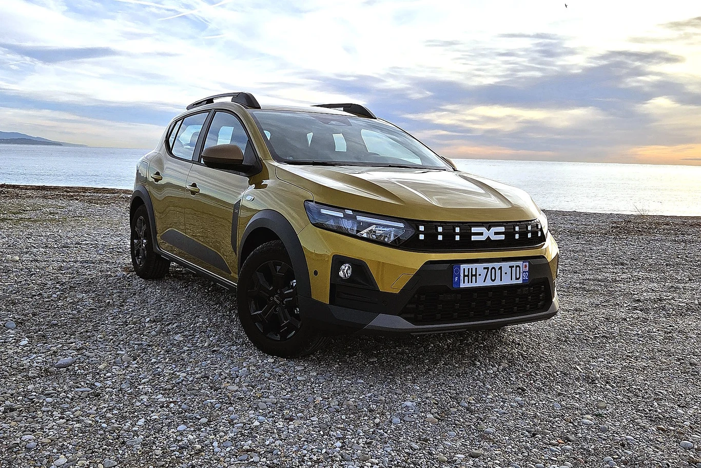 Żółty samochód osobowy Dacia stoi zaparkowany na kamienistej plaży nad morzem podczas zachodu słońca, widoczne tablice rejestracyjne oraz nowoczesny design pojazdu.