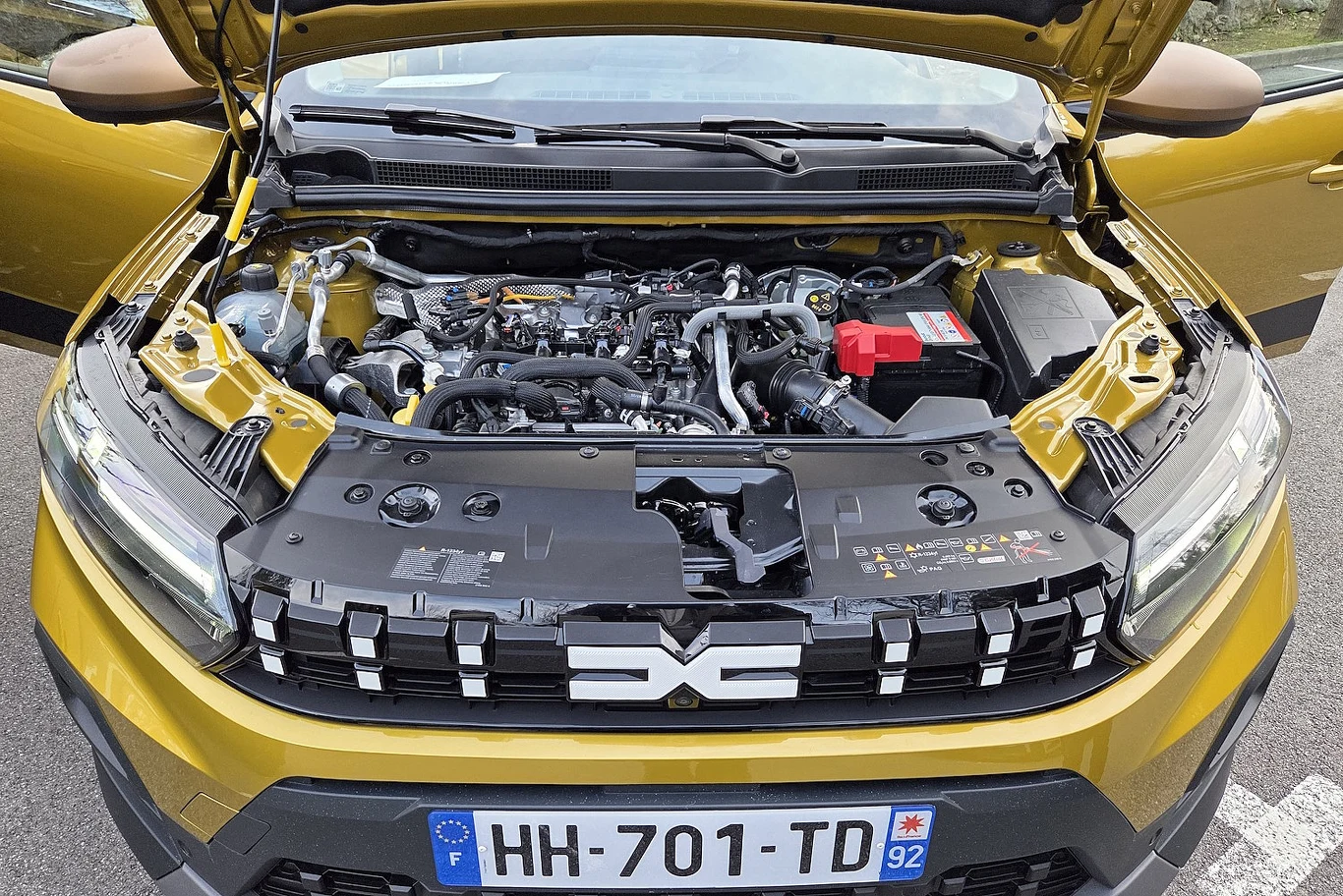Dacie Duster, Bigster, Sandero i Jogger otrzymały nowy podstawowy silnik - trzycylindrowy 1,2 l o mocy 120 KM i 200 Nm Maska samochodu uniesiona, widoczny nowoczesny silnik wraz z przewodami oraz baterią, na dole część grilla z logo producenta Dacia, numery rejestracyjne pojazdu wskazujące na rejestrację we Francji.
