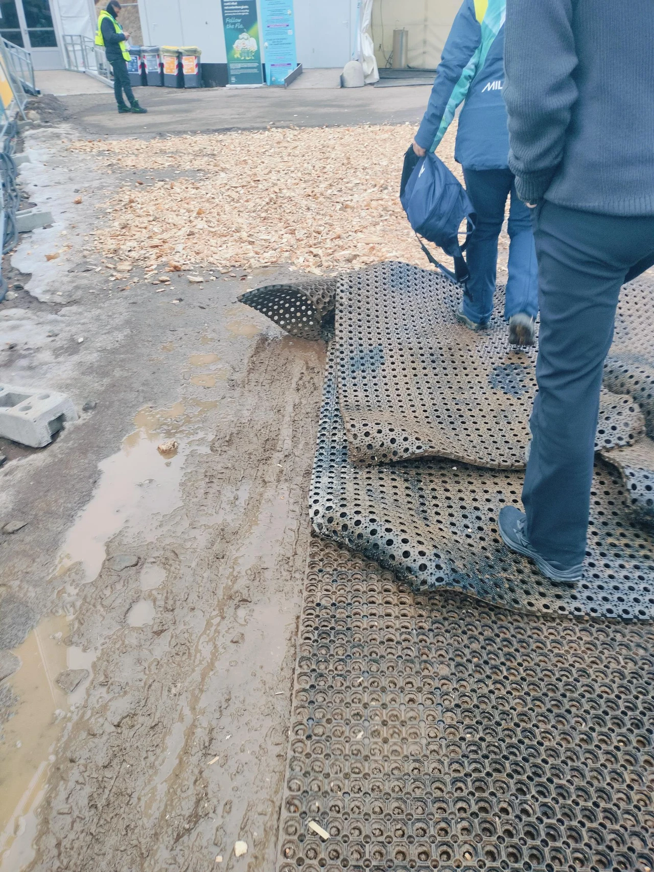 Tak wyglądała droga na skocznię w Predazzo. W wielu miejscach było podobnie Grupa osób idzie po ścieżce wyłożonej gumowymi matami ułożonymi na błotnistym terenie, w tle widoczni pracownicy w odblaskowych kamizelkach, plastikowe barierki i zabudowania tymczasowe.