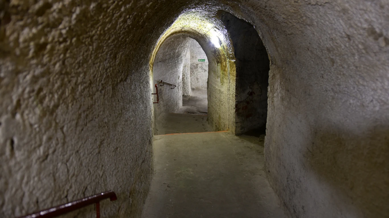 Kamienny tunel podziemny z oświetleniem na suficie, schodzący korytarz z balustradą po lewej stronie, wilgotne ściany i łuki, delikatnie oświetlone wejście w dalszej części korytarza.
