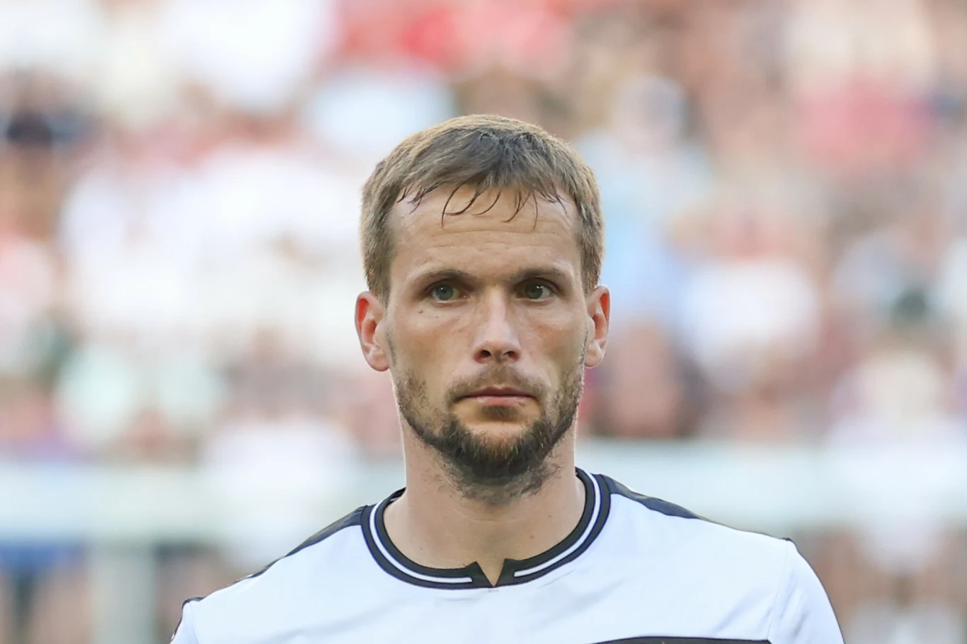 Mężczyzna z zarostem w sportowej koszulce podczas wydarzenia sportowego, w tle rozmyte sylwetki widzów lub kibiców.