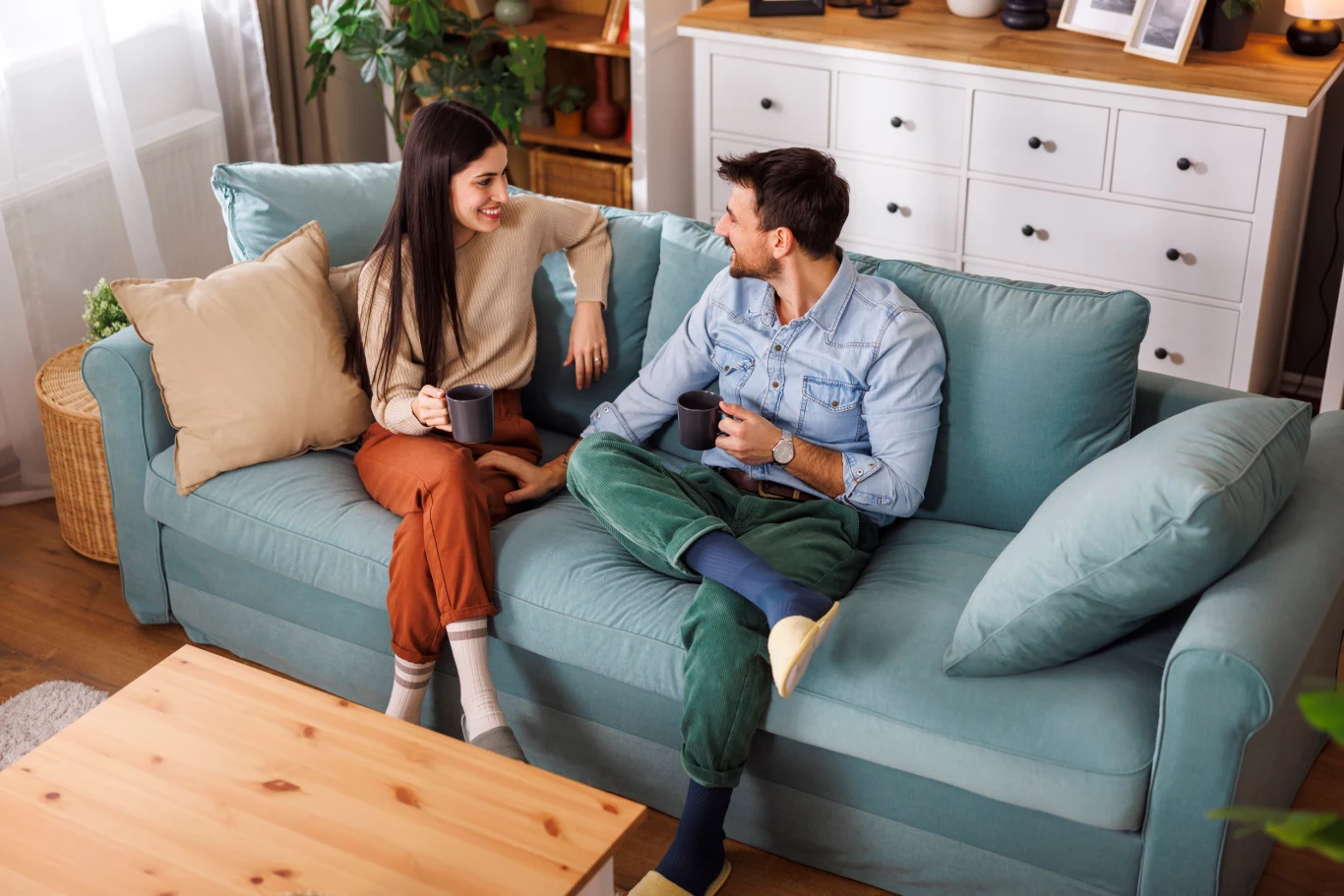 Dwoje młodych ludzi siedzi wygodnie na niebieskiej sofie w jasnym salonie, rozmawiając i uśmiechając się do siebie, trzymając w dłoniach kubki z napojem; atmosfera jest swobodna i przyjazna.