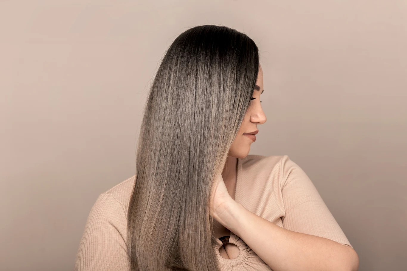 Ten zabieg świetnie zadziała zarówno na włosy proste jak i kręcone Kobieta z długimi, prostymi, gładkimi włosami w odcieniu ombre, ubrana w beżowy sweter, patrzy w bok i delikatnie dotyka szyi, tło jednolicie jasne.