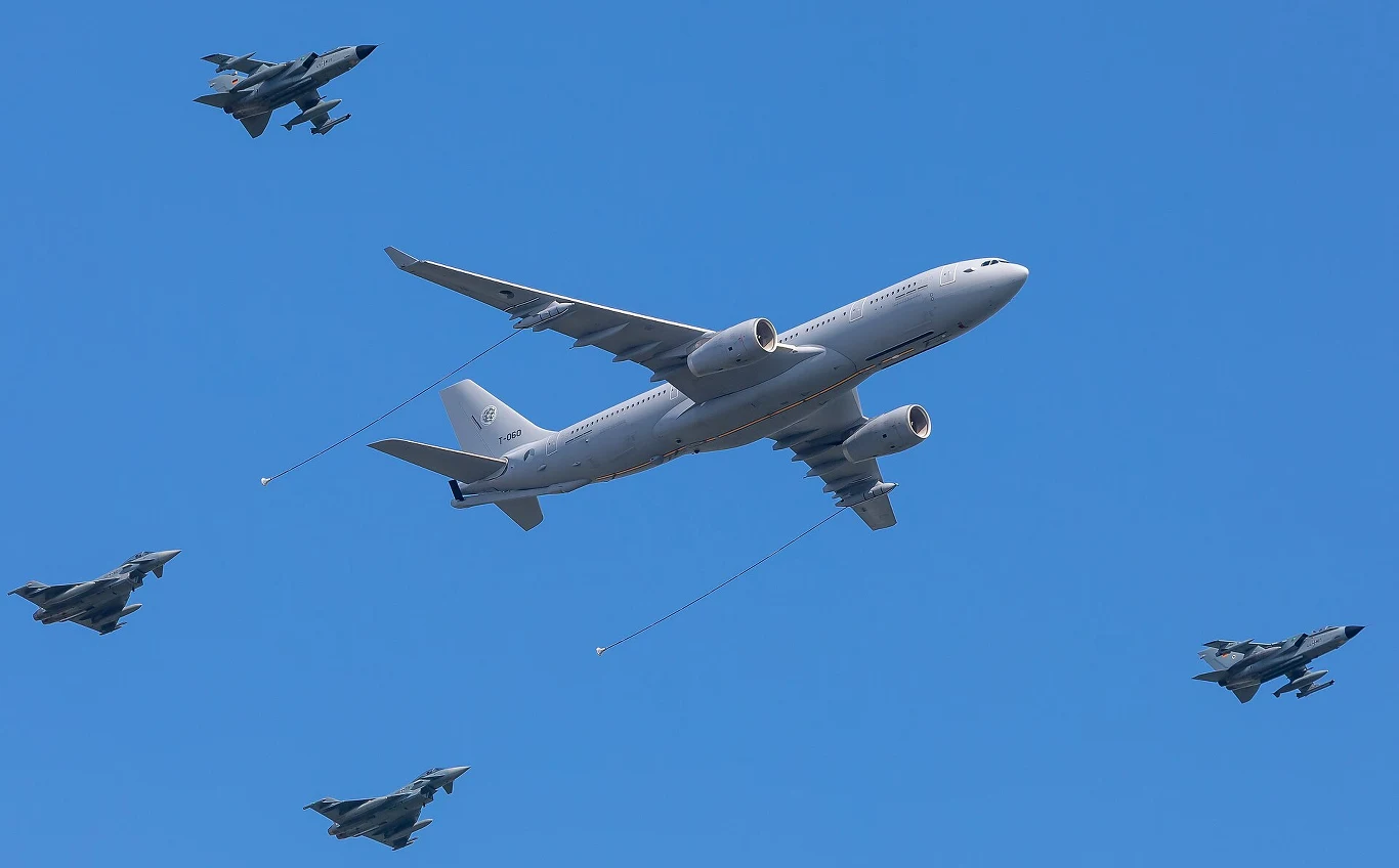 Duży samolot wojskowy w otoczeniu czterech myśliwców na tle czystego nieba, maszyny lecą w formacji synchronizując pozycje.