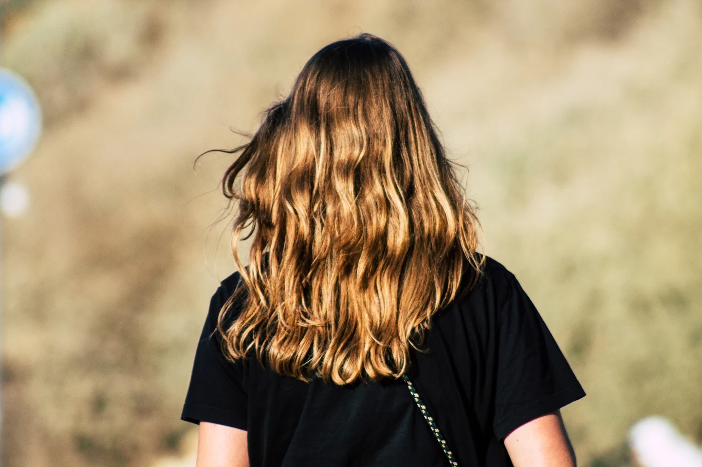 Osoba z długimi, falowanymi blond włosami ubrana w czarną koszulkę, widziana od tyłu, stoi na tle rozmytego, jasnego tła.