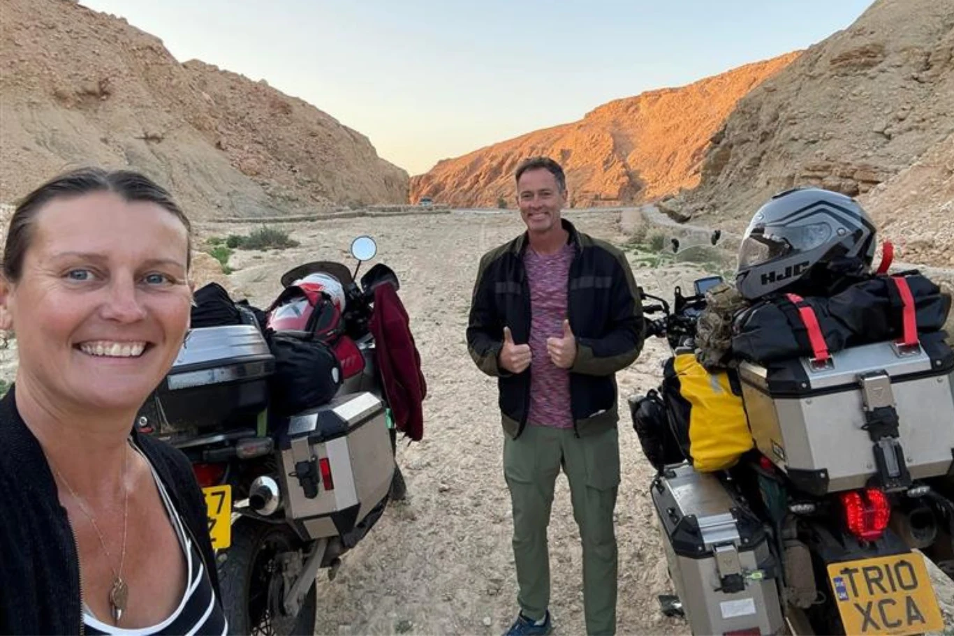 Para z Wielkiej Brytanii pojechała w podróż dookoła świata. Zostali zatrzymani w Iranie. / FB. Lindsay Foreman Uśmiechnięta kobieta i mężczyzna stoją przy dwóch motocyklach zapakowanych bagażami, otoczeni skalistym, pustynnym krajobrazem i oświetleni ciepłym światłem zachodzącego słońca.