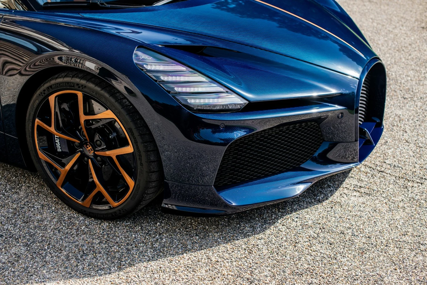 Przedni fragment niebieskiego sportowego samochodu z wyrazistym grillem oraz felgą w czarno-pomarańczowych kolorach, stojący na asfaltowej nawierzchni.