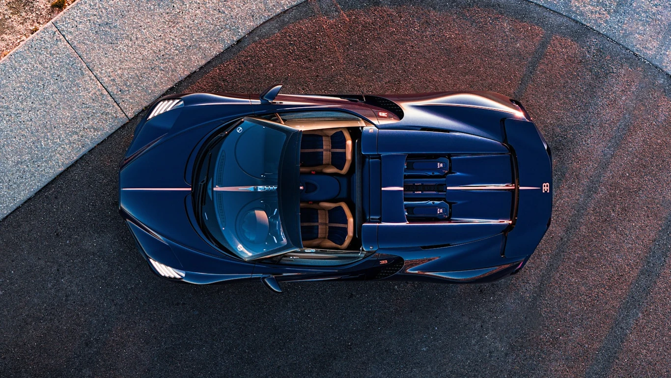 Luksusowy, sportowy samochód cabrio o ciemnogranatowym kolorze nadwozia zaparkowany na skraju wyłożonej kaflami nawierzchni, widziany z lotu ptaka. Wnętrze auta utrzymane w brązowej tonacji, z widocznymi siedzeniami oraz detalami silnika.