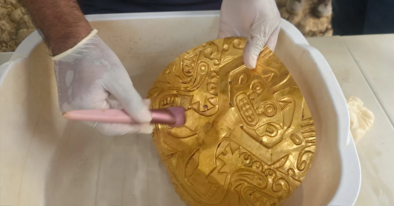 Prekolumbijska nekropolia elit w Panamie. Archeolodzy odkrywają ponad tysiącletni skarb Ręce w białych rękawiczkach czyszczą dużą złotą maskę ozdobioną reliefami przedstawiającymi geometryczne i zwierzęce motywy, maska znajduje się w pojemniku.