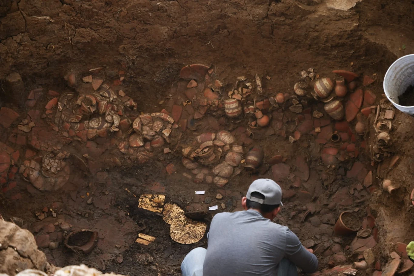 Archeolodzy odkryli skarby sprzed 1000 lat. Nawet dziś robią wrażenie Archeolog w szarej czapce obserwuje wykopalisko, w którym widoczne są stare gliniane naczynia oraz fragmenty złotych przedmiotów ułożone w ziemi.