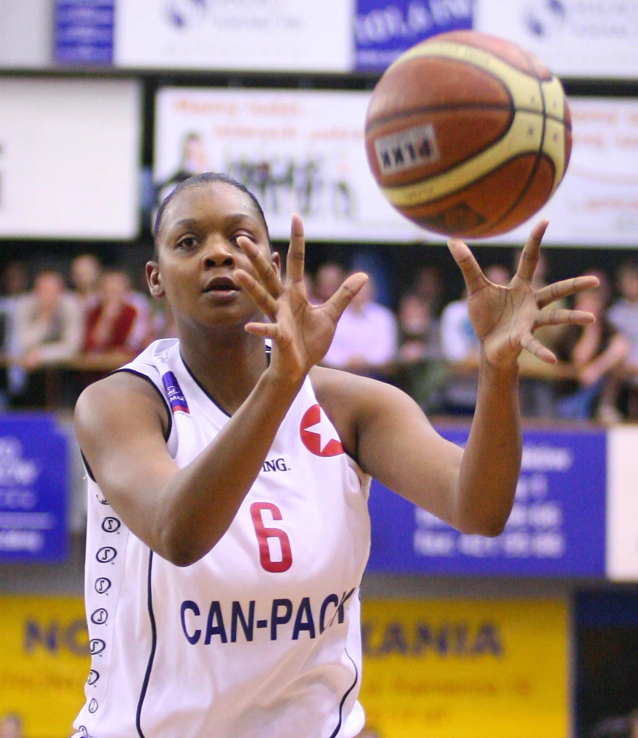 Koszykarka w białym stroju z numerem 6 łapie piłkę do koszykówki podczas meczu, w tle widownia i tablice reklamowe.