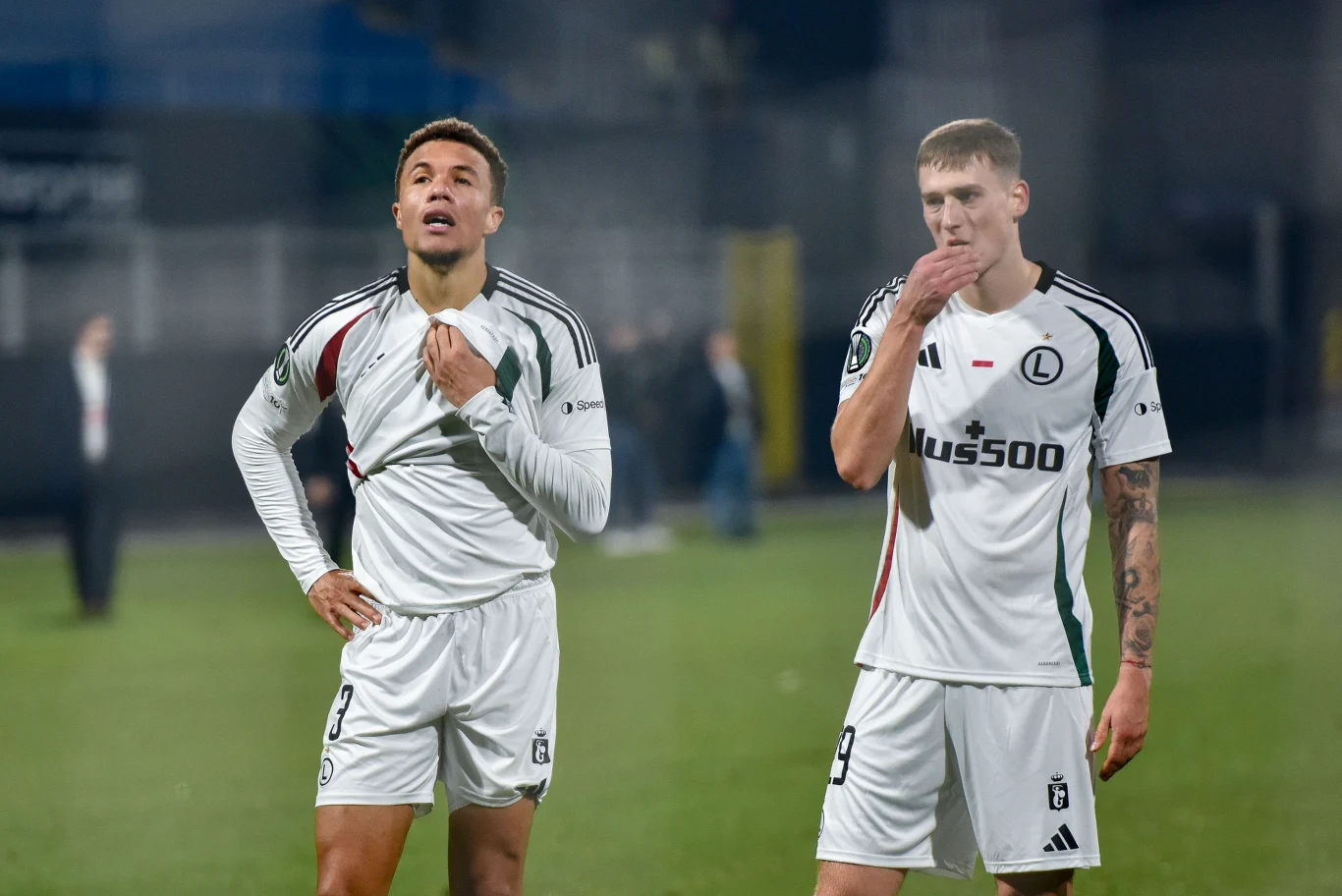 Dwóch piłkarzy w białych strojach klubowych Legii Warszawa stoi na murawie, obaj sprawiają wrażenie zamyślonych lub zawiedzionych, w tle rozmazana sylwetka innej osoby oraz fragmenty trybun.