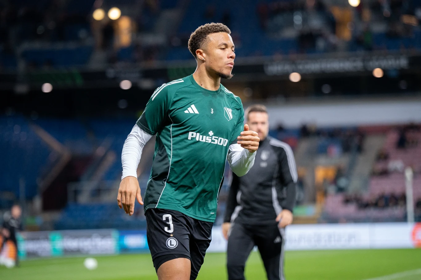 Steve Kapuadi Młody piłkarz w zielonej koszulce drużyny Legii Warszawa podczas rozgrzewki na murawie stadionu, w tle rozmyte postacie i trybuny.