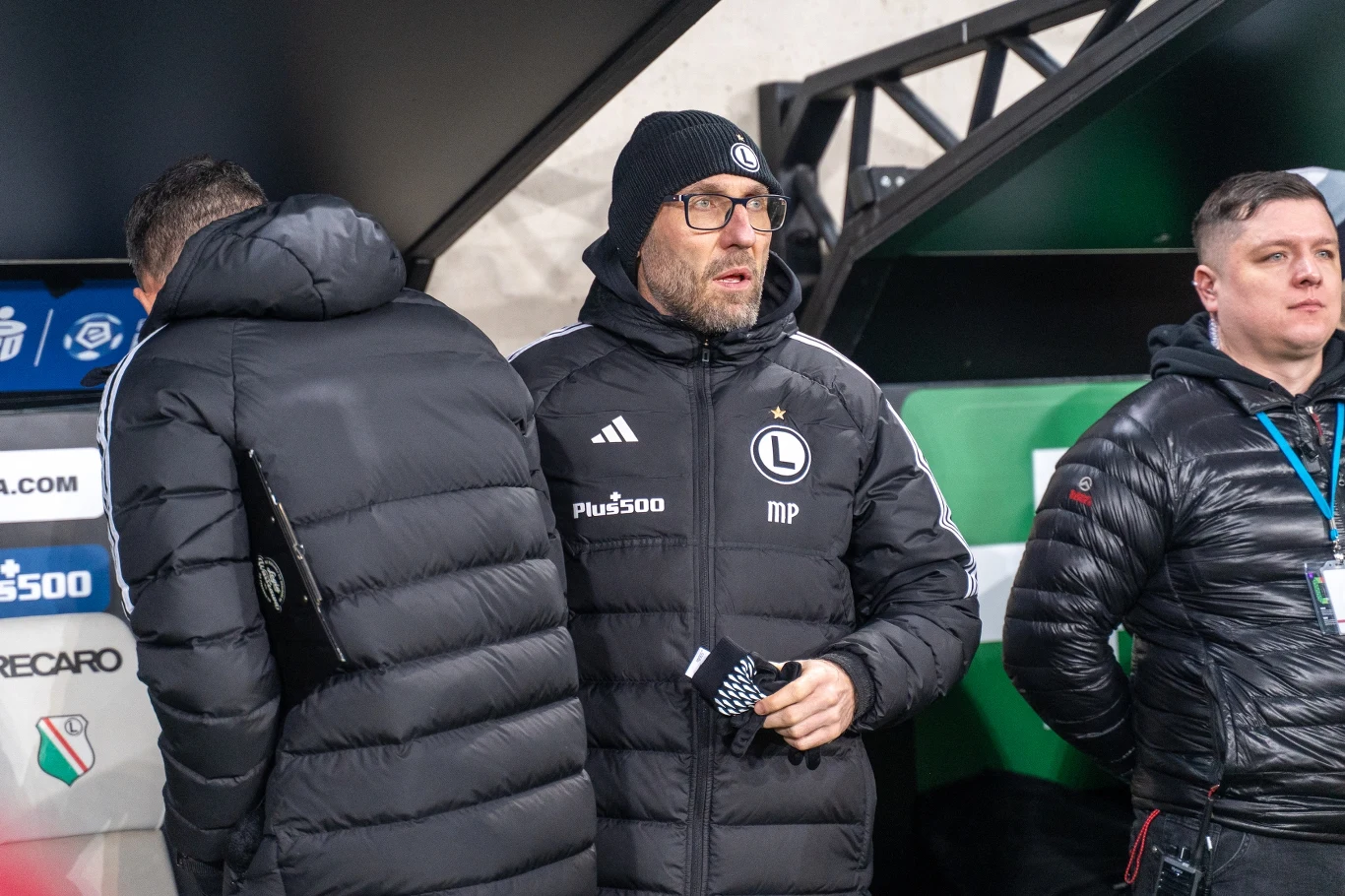 Trener ubrany w zimową kurtkę i czapkę z logo Legii Warszawa stoi obok innego mężczyzny w kurtce sportowej oraz ochroniarza z identyfikatorem, w tle ławka rezerwowych z widocznym logo klubu.
