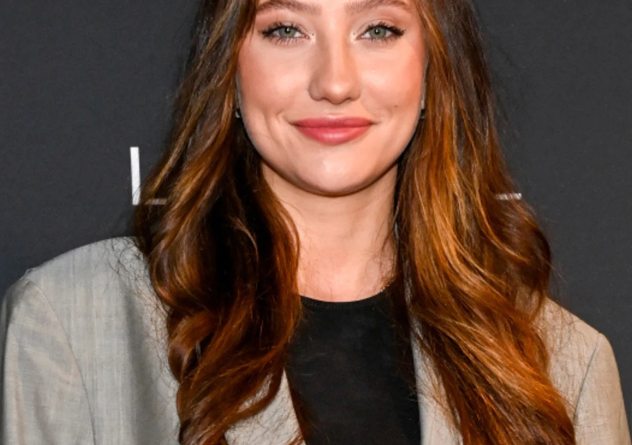 Klaudia Nieścior z "Top model" Młoda kobieta z długimi, falowanymi włosami w odcieniach brązu i rudego, uśmiechnięta, ubrana w szarą marynarkę oraz czarną bluzkę, stojąca na tle ciemnego, jednolitego tła.