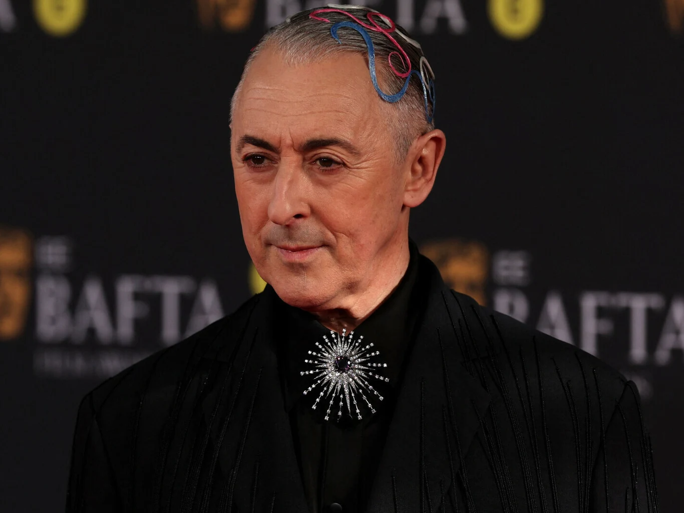 Alan Cumming podczas 79. ceremonii rozdania nagród BAFTA Mężczyzna w eleganckim, ciemnym garniturze z oryginalną ozdobą przypinaną do klapy, z kolorowymi elementami na włosach, pozuje na tle tablicy z logo BAFTA.