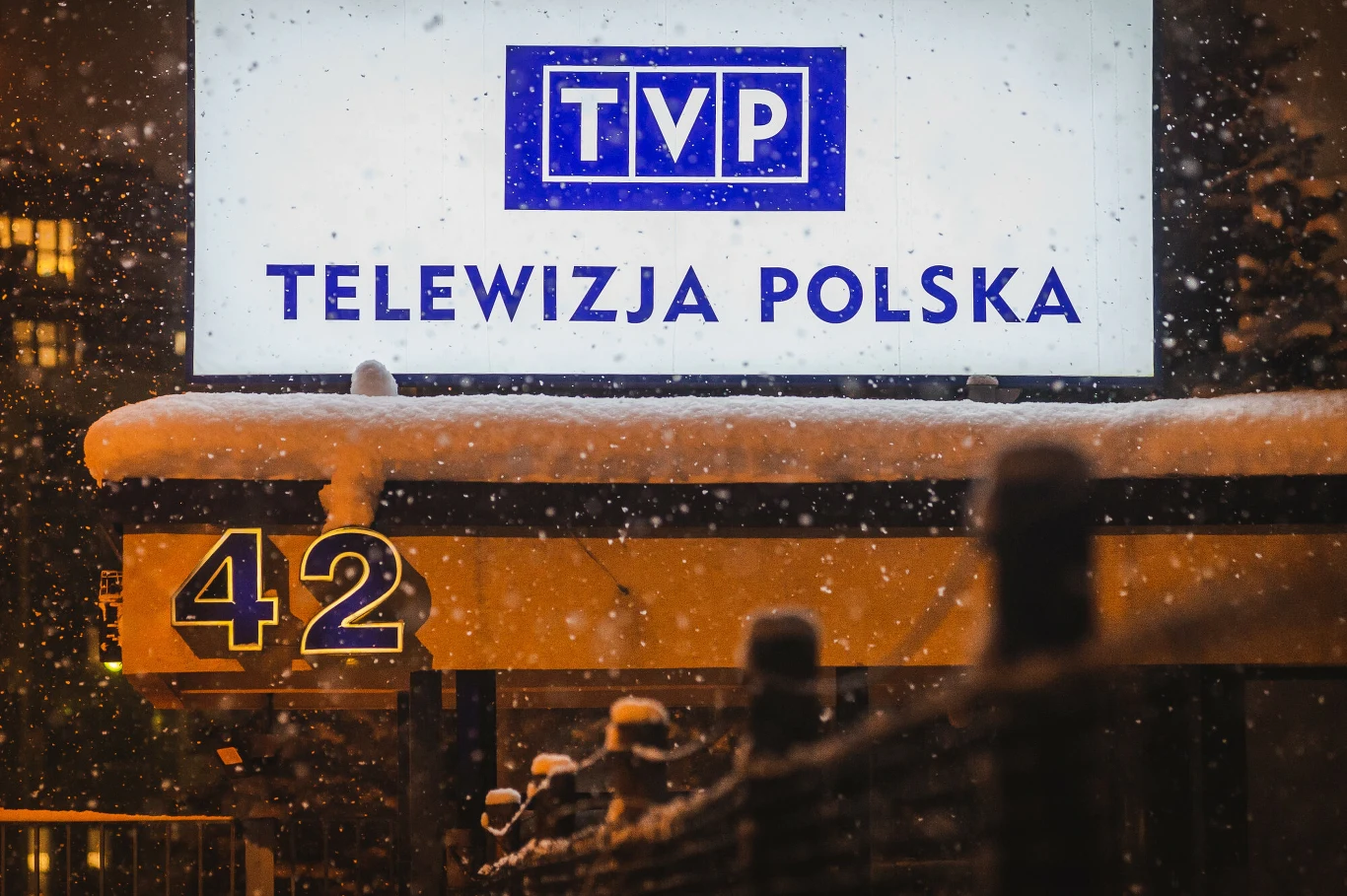 Neonowy szyld z napisem TVP Telewizja Polska oraz numerem 42, pokryty warstwą śniegu, w otoczeniu wieczornej scenerii z widocznymi opadami śniegu.