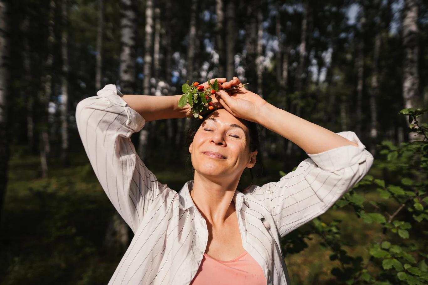 Kobieta w jasnej koszuli z zamkniętymi oczami uśmiecha się, trzymając na głowie wieniec z liści i jagód, stojąc w słonecznym lesie brzozowym.