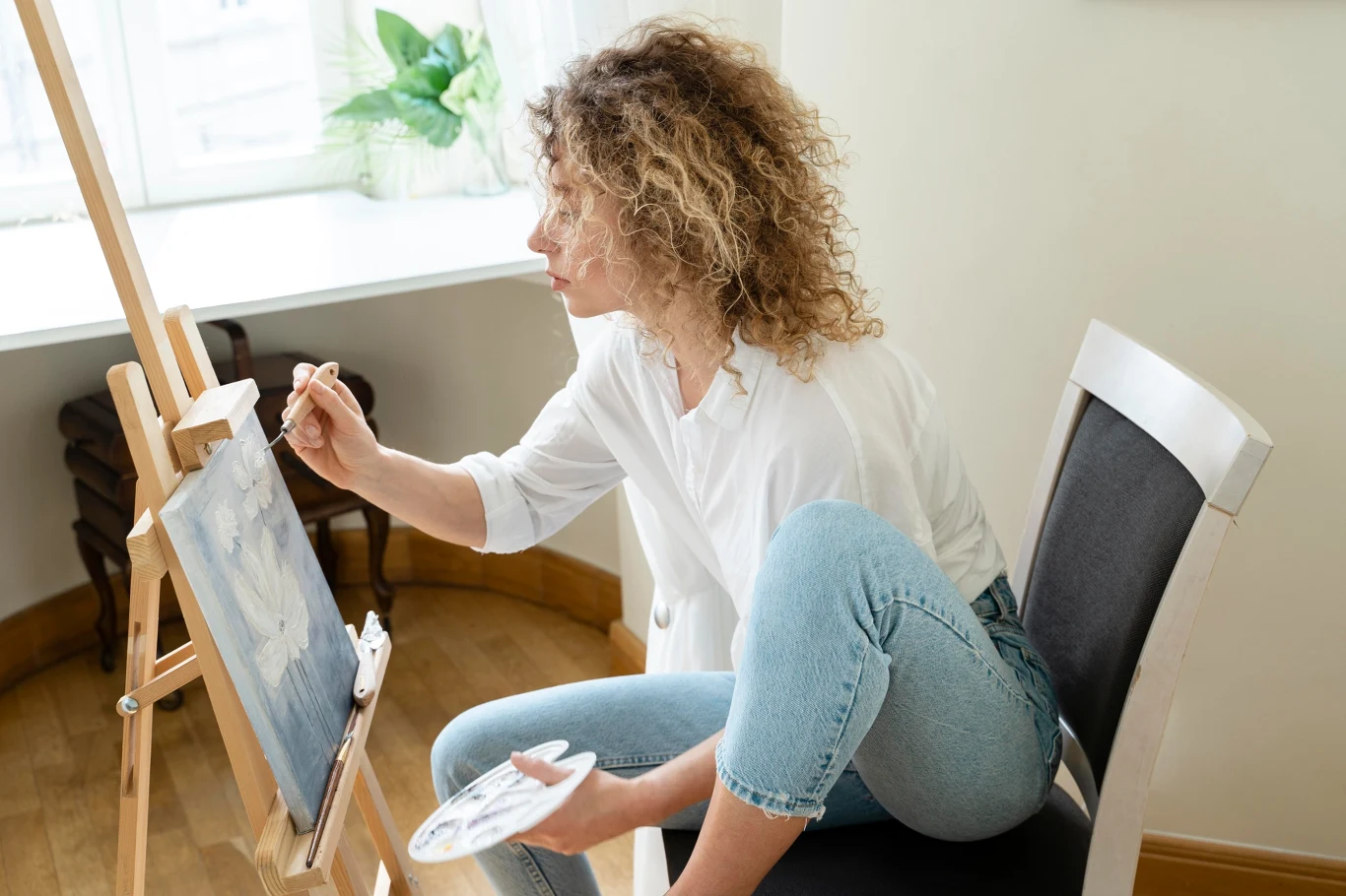Kobieta z kręconymi włosami siedzi na krześle przy sztaludze i maluje obraz farbami na płótnie w jasnym, domowym wnętrzu z rośliną w tle.