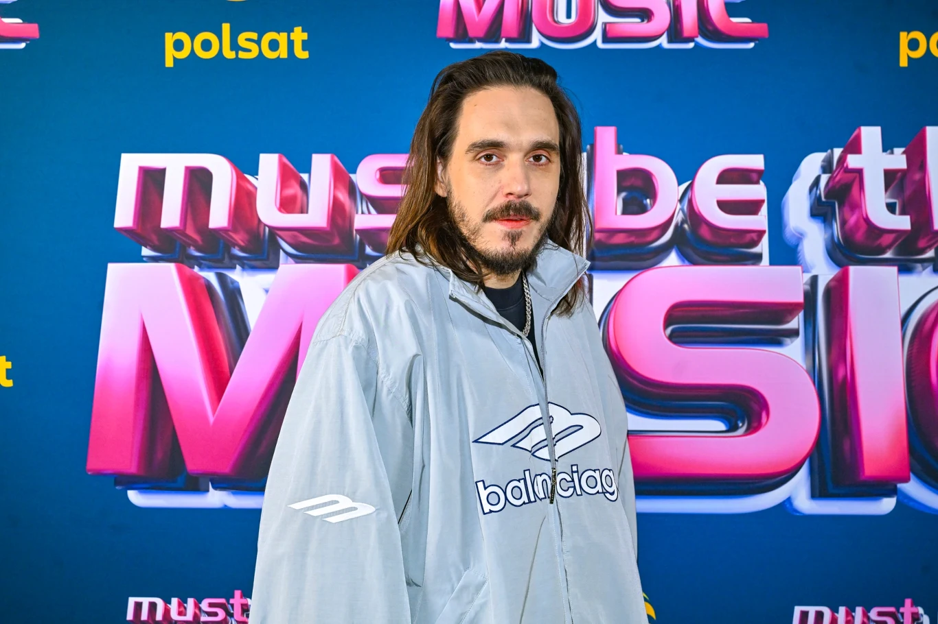 Mężczyzna z długimi włosami ubrany w jasną kurtkę z logo Balenciaga stoi na tle ścianki promującej program muzyczny Must Be The Music z widocznymi logotypami Polsatu.