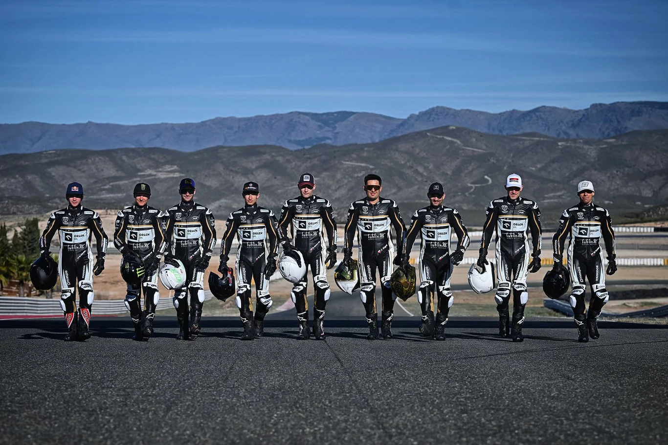 Grupa ośmiu motocyklistów w profesjonalnych kombinezonach, idących równym krokiem po torze wyścigowym z kaskami w rękach, w tle góry i niebieskie niebo.