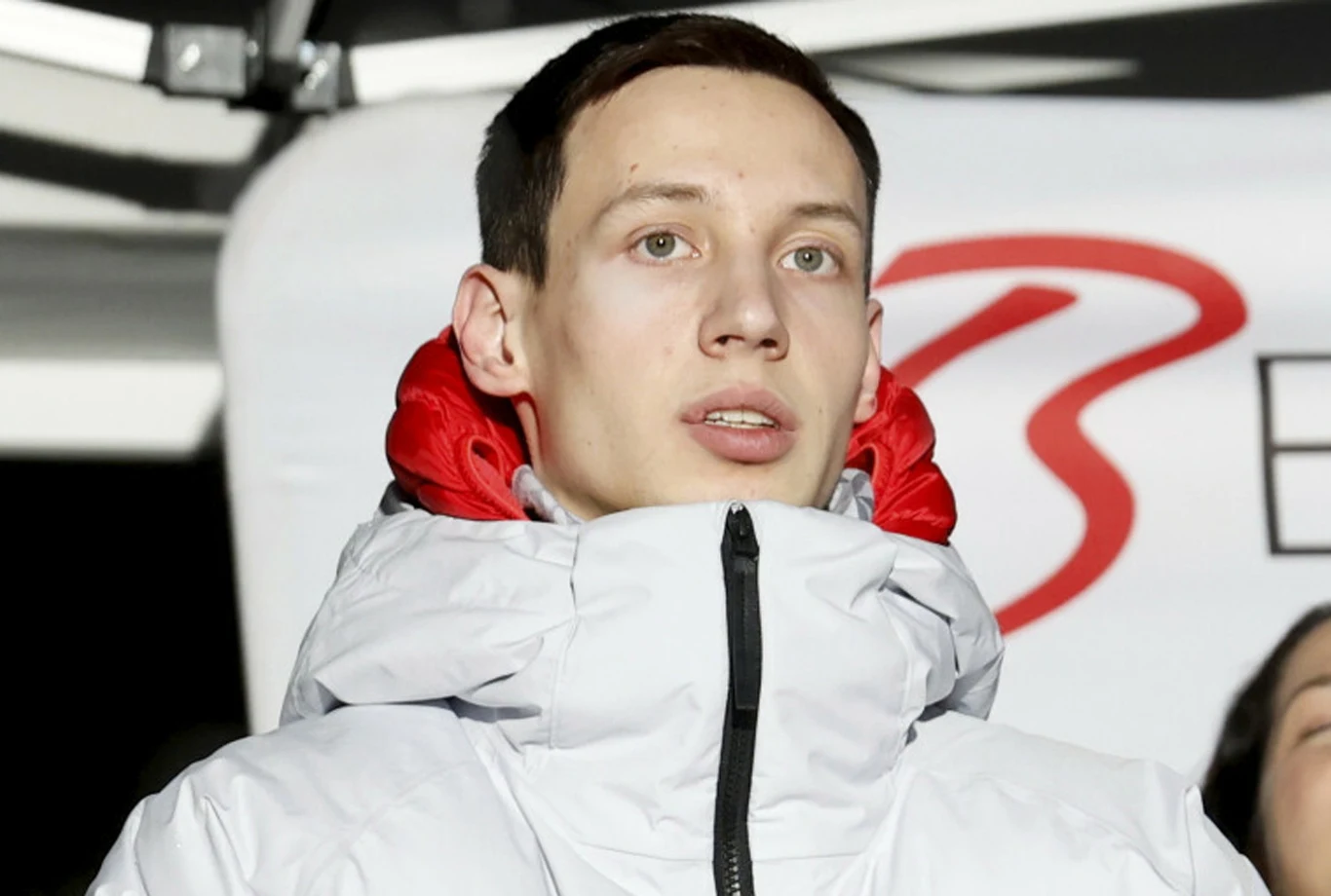 Kacper Tomasiak Młody mężczyzna w jasnej kurtce z wysokim kołnierzem, czerwonym podszyciem i ciemnymi włosami stoi na tle rozmytego znaku z literą B.