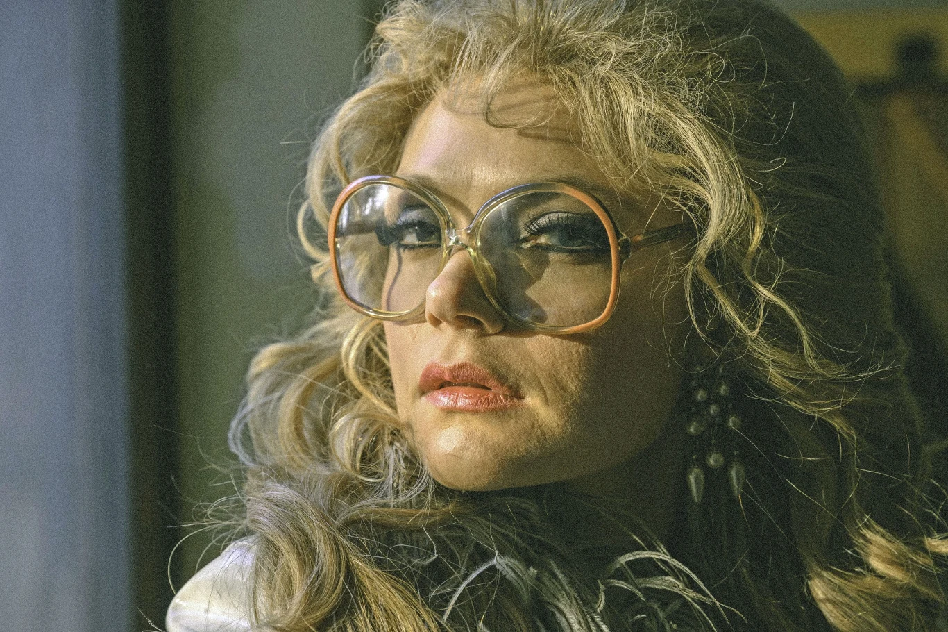 Kobieta z blond, falowanymi włosami w dużych, okrągłych okularach o grubych oprawkach, z makijażem i srebrzystym elementem ubioru, patrząca w bok, o wyrazistym wyrazie twarzy.