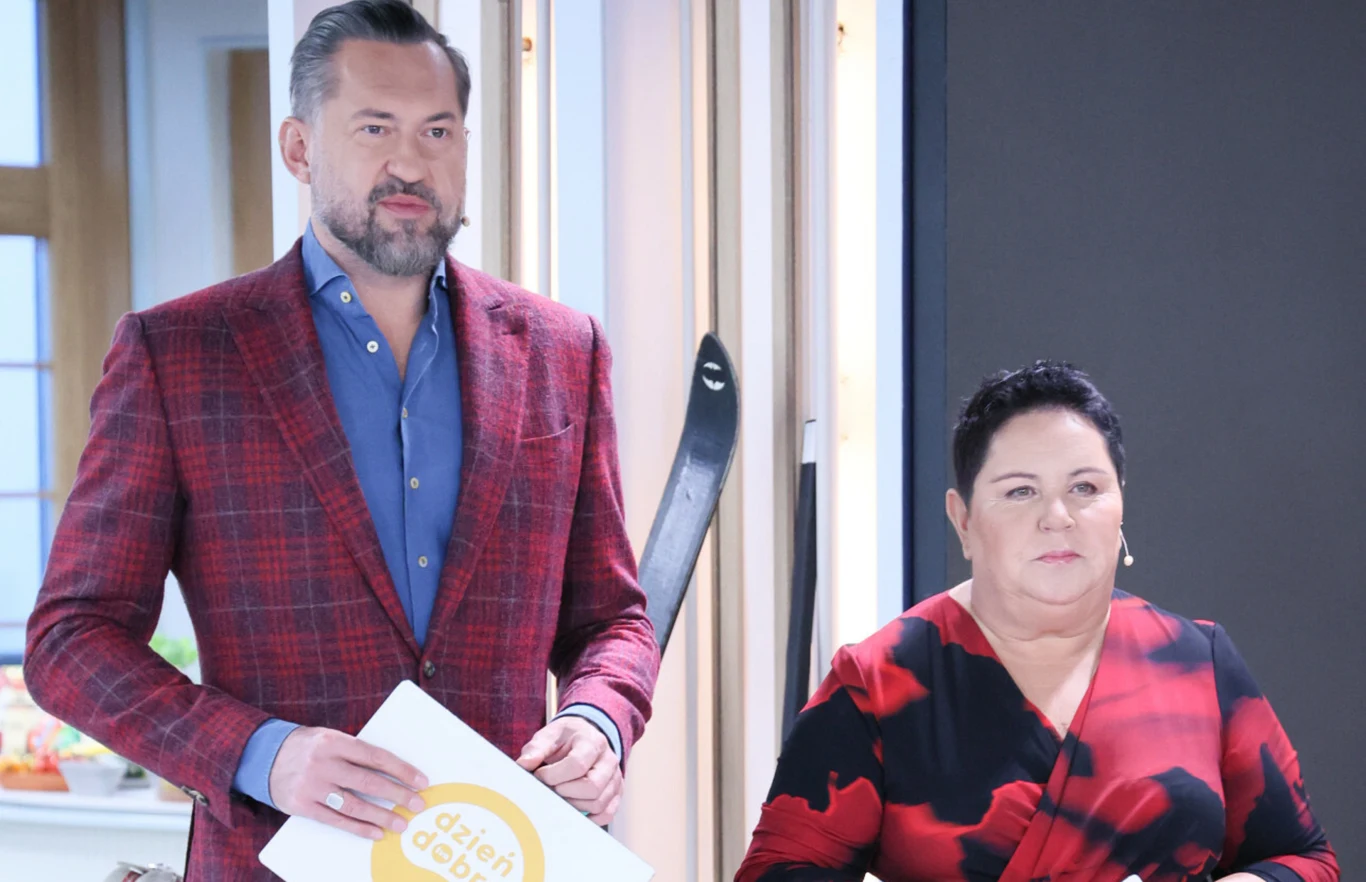 Marcin Prokop, Dorota Wellman Dwójka prowadzących w studiu telewizyjnym, mężczyzna w czerwonej marynarce trzymający kartkę z napisem 'Dzień Dobry', obok stoi kobieta w czerwono-czarnej sukni, tło stanowią jasne wnętrza i narty oparte o ścianę.