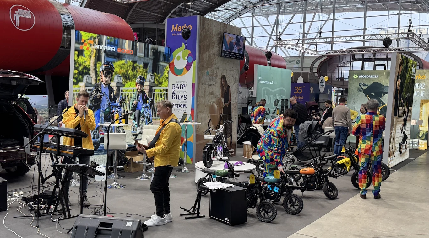 Występ zespołu muzycznego przed publicznością w przestrzeni targowej, w otoczeniu stoisk z rowerami dziecięcymi oraz osobami w kolorowych strojach przypominających kostiumy klaunów. W tle widoczne bannery reklamowe oraz nowoczesna architektura hali.