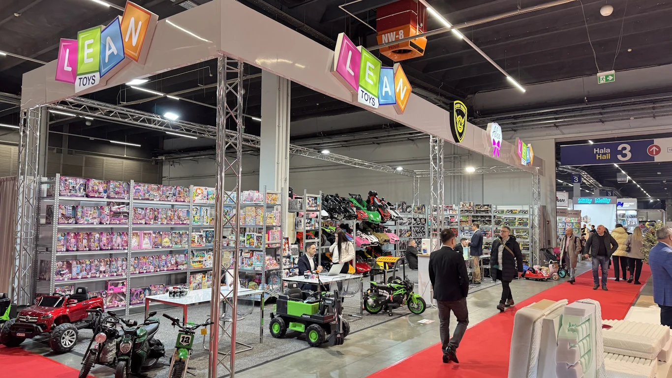 Stoisko targowe firmy z zabawkami z wyeksponowanymi półkami pełnymi różnorodnych produktów dziecięcych, samochodami elektrycznymi, motocyklami na akumulator oraz licznymi kolorowymi opakowaniami. Po czerwonym dywanie przechodzą zwiedzający i osoby obsł...