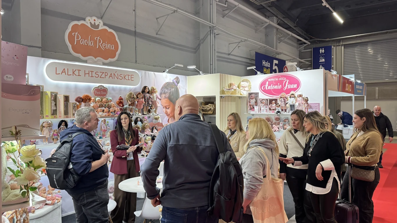 Stoisko targowe z hiszpańskimi lalkami otoczone przez grupę osób, które oglądają ekspozycję i rozmawiają między sobą. Kolorowe stoiska prezentujące lalki, zabawki oraz banery reklamowe marek. Centralnym punktem są rozmowy uczestników i zauważalna atmos...