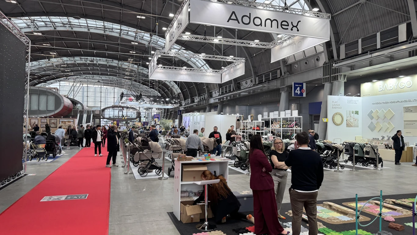 Hala targowa z licznymi stoiskami firm, w tym widocznym szyldem Adamex, czerwonym dywanem prowadzącym przez centralną część oraz grupami ludzi rozmawiających i oglądających produkty, głównie wózki dziecięce.