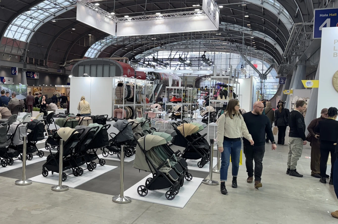 Stoisko targowe z dużą liczbą wózków dziecięcych ustawionych w rzędach, otoczone barierkami. W tle widoczna ekspozycja innych produktów dla dzieci oraz przechodzące osoby zwiedzające hale targową o nowoczesnej konstrukcji.