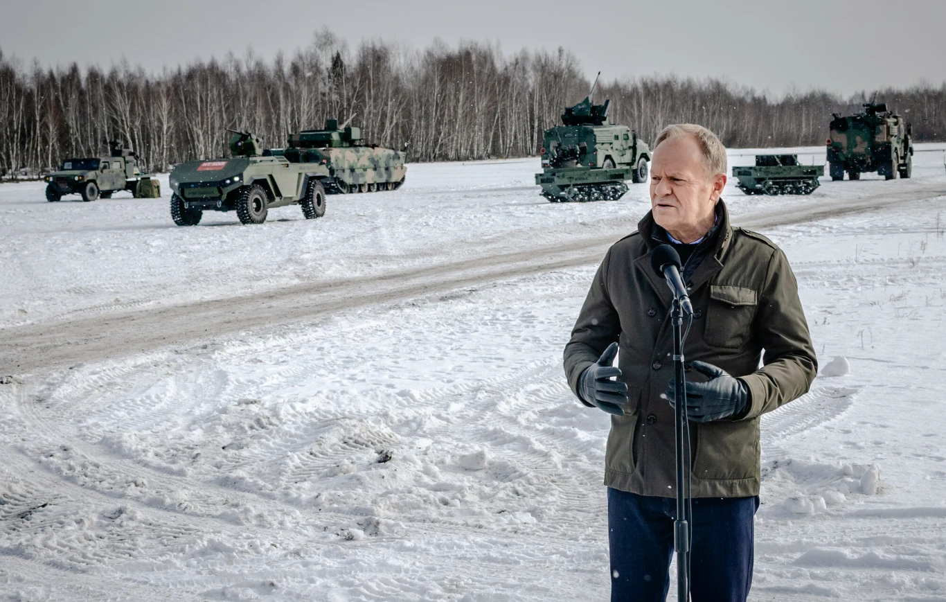 Donald Tusk spotkał się z szefem rządu Norwegii Jonasem Gahrem Storem. Zdjęcie ilustracyjne. Mężczyzna przemawia do mikrofonu na tle wojskowych pojazdów opancerzonych ustawionych na zaśnieżonym terenie, w oddali widoczne drzewa bez liści i zimowy krajobraz.