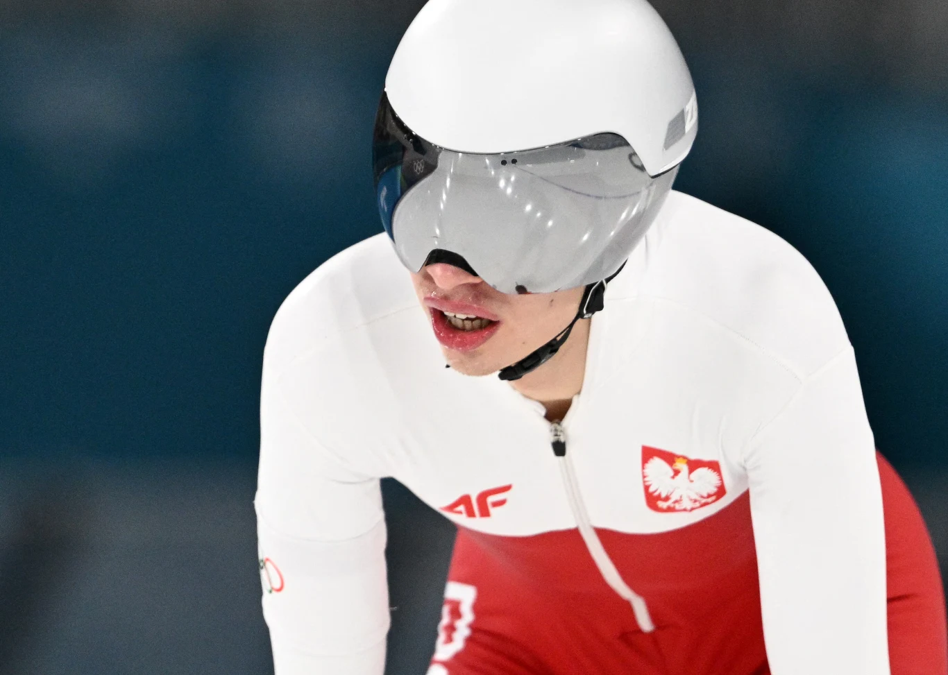 Zawodnik w biało-czerwonym stroju reprezentacji Polski do łyżwiarstwa szybkiego podczas jazdy na torze, w aerodynamicznym kasku z przyciemnianą szybą.