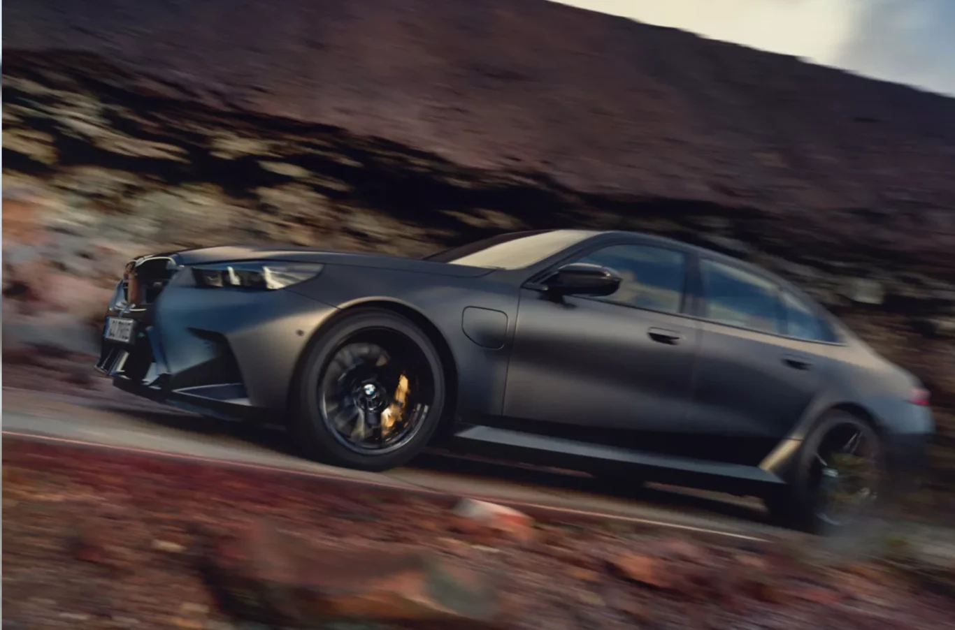 Egzemplarz Urbańskiego to wersja z układem hybrydowym typu plug-in, łącząca silnik benzynowy V8 o pojemności 4,4 litra z jednostką elektryczną. Czarny sportowy sedan jedzie w szybkim tempie po drodze, tło rozmyte z wyrazistą czarną karoserią w centrum kadru, za pojazdem widoczna skalista ściana.